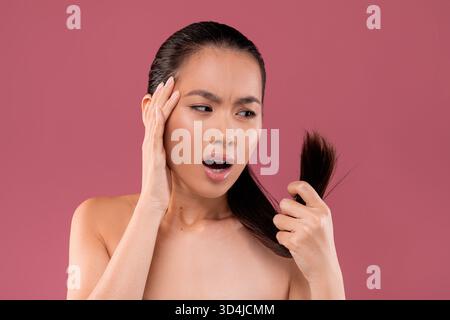 La giovane donna asiatica preoccupata con la coda di cavallo ispeziona i suoi capelli per le estremità spaccate su sfondo viola Foto Stock