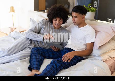 Puntando verso tablet diversi partner maschi che si rilassano sul letto nella moderna camera da letto, con tavolino in legno Foto Stock