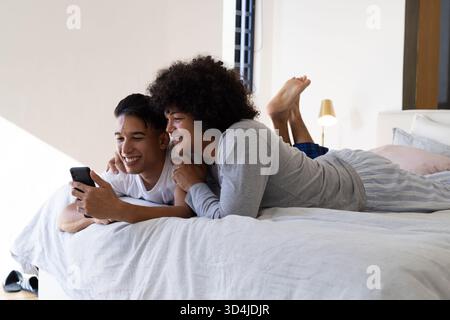 Sdraiati diversi partner maschili che si coccolano sul letto di casa tenendo in mano lo smartphone con cuscini e lampada color pastello Foto Stock