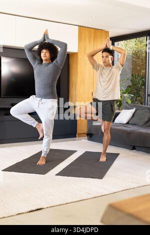 Praticare diversi amici maschi in posa sugli alberi nel soggiorno moderno, con tappetini neri per lo yoga Foto Stock