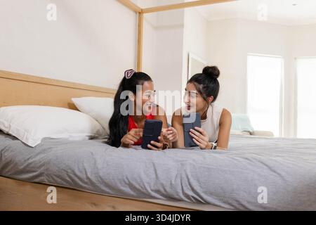 Sdraiate diverse amiche che chiacchierano su smartphone a letto in una camera da letto luminosa, con cornice a baldacchino Foto Stock
