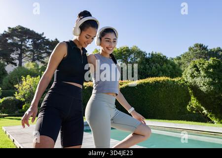 Equilibrare e piegare diverse amiche sul ponte a bordo piscina indossando cuffie e smartwatch Foto Stock