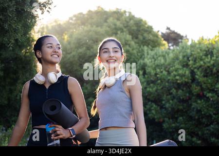 Diverse amiche che camminano nel parco portando con sé stuoie da yoga, con cuffie e bottiglia d'acqua Foto Stock