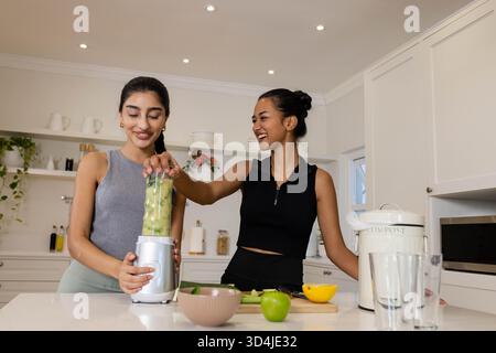 Mescola diversi amici di sesso femminile mescolando frullati verdi all'isola della cucina, con frullatore e frutta Foto Stock