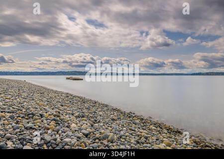 Una fotografia minimalista a lunga esposizione di una spiaggia rocciosa, che mostra acqua liscia e calma e nuvole striate sul Puget Sound, con una piccola piattaforma all'interno Foto Stock