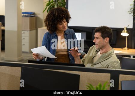 Colleghi diversi che discutono documenti con la penna alla scrivania dell'armadio in un ufficio moderno, con monitor Foto Stock