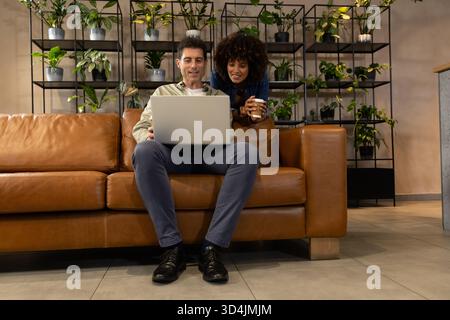 Lavorare con colleghi diversi utilizzando un computer portatile tenendo la tazza del caffè sul divano nell'area salotto dell'ufficio Foto Stock