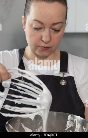 Un esperto pasticcere si concentra sulla preparazione della meringa alla perfezione in una cucina moderna. La scena cattura l'attento processo di creazione di un dolce regalo, mostrando dedizione e competenza. Foto Stock