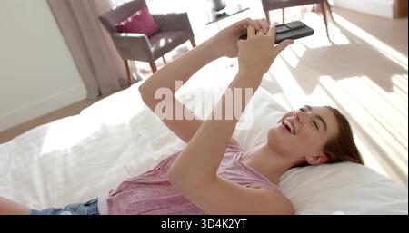 Donna che ride indossa un top rosa e pantaloncini in denim che reggono il telefono sopra il piumone bianco in camera da letto Foto Stock