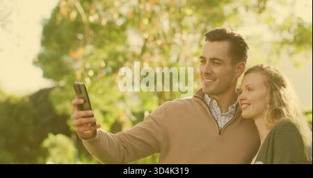 Coppia sorridente che posa per selfie al parco, uomo che tiene il telefono, indossa un maglione abbronzante, cardigan scuro Foto Stock