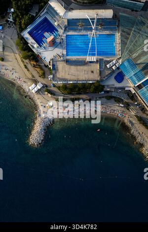 Vista aerea di una spiaggia vivace brulicante di bagnanti accanto a un moderno complesso di piscine azzurre, che contrasta nettamente con il mare blu profondo, Foto Stock
