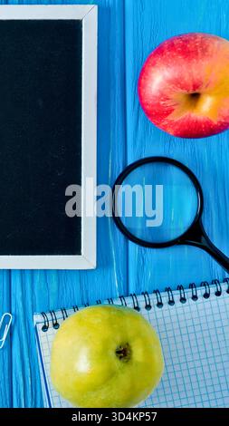 Una tavoletta, una lente di ingrandimento, un quaderno e due frutti sono disposti su una superficie in legno blu brillante, suggerendo un tema di studio o di ricerca. Foto Stock