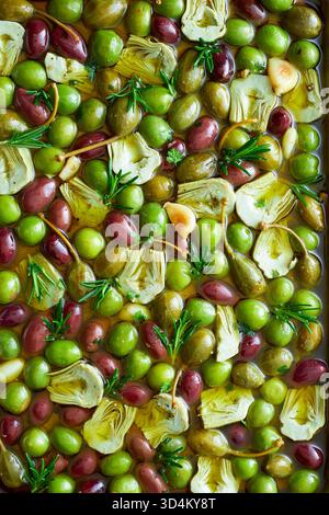 Fotografia gastronomica di alta qualità di antipasti mediterranei: Un mix di olive verdi e nere, cuori di carciofo, aglio, rosmarino e frutti di cappero mari Foto Stock