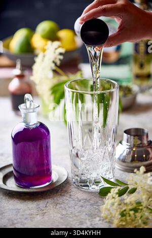 Un primo piano d'azione di una mano che versa un liqour chiaro in uno shaker di cocktail di cristallo. Una bottiglia in stile vintage riempita con flowe di piselli farfalla viola Foto Stock