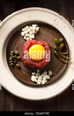 deposizione piatta di tartare di manzo crudo da vicino in un piatto con diversi tipi di condimenti, un tuorlo d'uovo crudo, capperi, frutti di cappero, cipolle tritate, su un legno Foto Stock