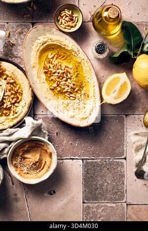 stesa piatta di un piatto ovale con hummus decorato con pinoli e una ciotola di pasta di miso a lato con limoni freschi e olio d'oliva sul piano del tavolo piastrellato Foto Stock