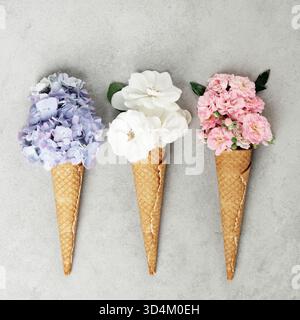 Trio creativo di coni di waffle pieni di ortensie, rose bianche e garofani rosa disposti su una superficie grigia. Foto Stock