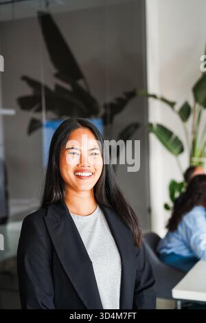 Giovane donna d'affari asiatica sorridente sulla macchina fotografica all'interno dell'ufficio aziendale - concetto imprenditoriale femminile - Focus on Face - foto verticale Foto Stock