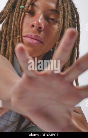 Giovane con dreadlock che si allungano il braccio e la mano aperta verso lo spettatore in studio Foto Stock