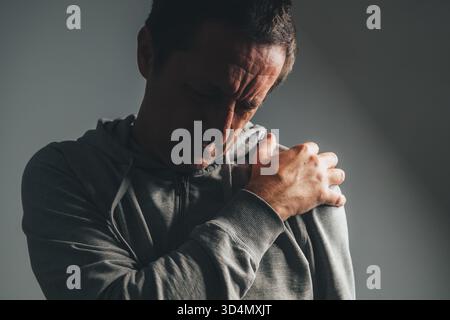 Uomo maturo con una luce chiave bassa che tiene la spalla in gravi dolori, concetto di lesioni, tensione muscolare o problemi cronici alle articolazioni. Messa a fuoco selettiva. Foto Stock