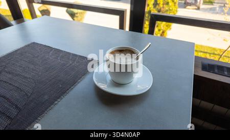 Tazza di cappuccino sul tavolo vicino alla finestra in una caffetteria soleggiata, stile di vita semplice e concetto di relax mattutino Foto Stock