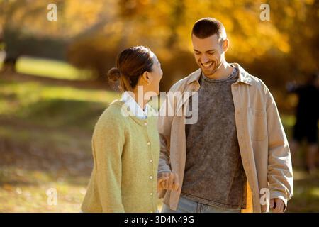 Una gioiosa donna coreana e un allegro uomo caucasico camminano mano nella mano attraverso un vivace parco autunnale, circondato da un fogliame colorato. Foto Stock