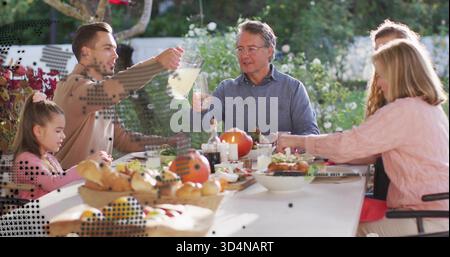 Versare l'uomo nel maglione abbronzante sollevando la caraffa mentre la famiglia è seduta sul patio del giardino, con cestino del pane Foto Stock