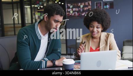 Discutendo di colleghi che si rivolgono a un notebook argentato nella sala d'ufficio, un blazer abbronzante e un cardigan verde Foto Stock