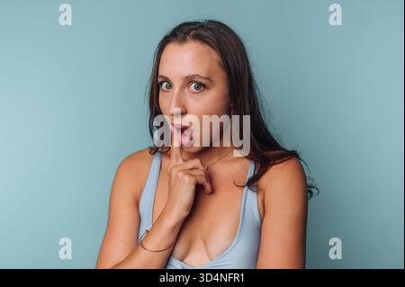 Una giovane donna si erge su uno sfondo azzurro, esprimendo sorpresa con gli occhi larghi e il dito alzato alle labbra. Ha i capelli lunghi e i vestiti Foto Stock