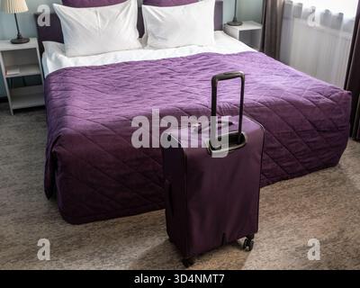 valigia viola e letto matrimoniale con biancheria bianca e copriletto viola, con cuscini nel piccolo soggiorno Foto Stock