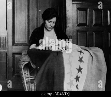 Ritratto a mezza lunghezza di Alice Paul, seduta e rivolta leggermente a destra, cucendo una bandiera a suffragio. La fotografia cattura Paul, una delle principali suffragiste e stratega americane del National Woman’s Party, impegnato in attivismo per il diritto di voto delle donne all’inizio del XX secolo. Foto Stock