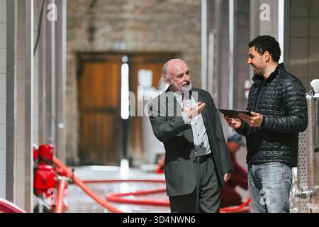 Due uomini sono impegnati in una conversazione all'interno di un'azienda vinicola, discutendo delle operazioni e ispezionando le attrezzature in uno spazio luminoso. Foto Stock