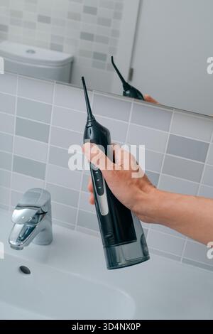 una vista ravvicinata di un uomo che solleva un irrigatore orale davanti allo specchio in un bagno interno piastrellato, con un moderno rubinetto cromato Foto Stock