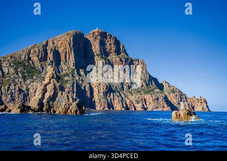Cap rosso con scogliere rosse scoscese e torre genovese nel Golfo di Porto, vista dal mare sotto il cielo azzurro e la luce del sole Foto Stock