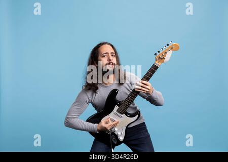Giovane chitarrista maschile con capelli lunghi che si esibisce con passione su una chitarra elettrica nera in una posa dinamica. Un musicista energico trasuda vibrazioni rock star, gioia ed entusiasmo in uno studio di registrazione. Foto Stock