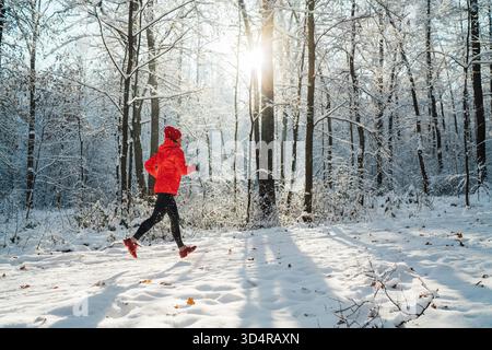 Trail runner donna vestita con una giacca antivento rossa brillante che fa jogging su un pittoresco sentiero innevato nella foresta durante una giornata soleggiata e gelida. Persone sportive attive e invernali Foto Stock