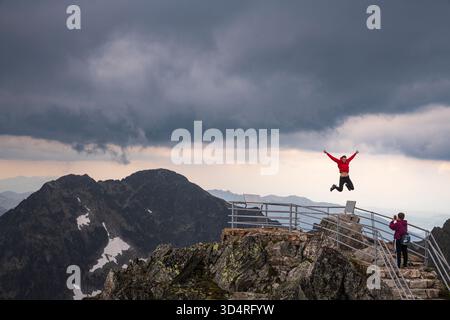 Il fotografo cattura i giovani che saltano per gioia con la vista della montagna degli alti Tatra presso la piattaforma di osservazione di Lomnický štít. Foto Stock