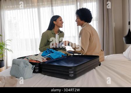 Imballare diverse coppie sedute sul letto in una camera da letto luminosa, smistare i vestiti in una valigia nera Foto Stock