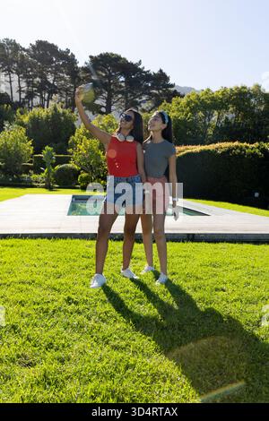 Diverse amiche che scattano selfie sul prato a bordo piscina, con smartphone e occhiali da sole Foto Stock