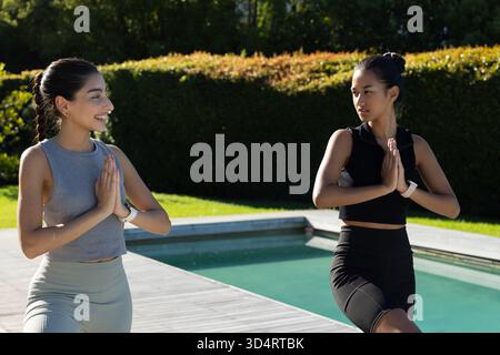 Diverse amiche che praticano la posa sull'albero sul ponte della piscina nel cortile posteriore, con smartwatch Foto Stock