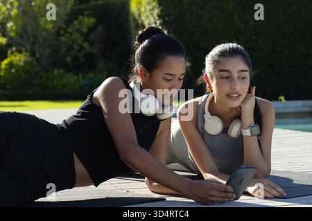 Sdraiate diverse amiche che controllano lo smartphone sui tappetini da ginnastica presso la piscina del cortile, con le cuffie Foto Stock