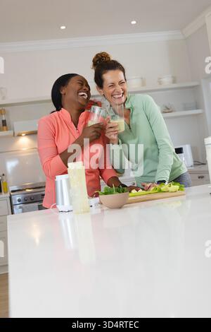 Ridendo le diverse amiche che brucano bicchieri di frullato verde all'isola della cucina, con frullatore Foto Stock