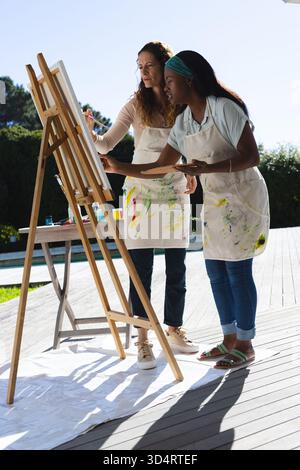 Diverse istruttrici e studentesse che dipingono sul ponte in legno a bordo piscina, con cavalletto e vasetti di pittura Foto Stock
