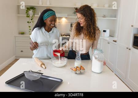Mescola diverse amiche che sbattono la pastella sull'isola della cucina, con recipiente per mescolare e dosatore Foto Stock