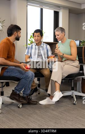 Chiacchierare con colleghi diversi che discutono del progetto in ufficio, con laptop, mela rossa e contenitore di frutta Foto Stock