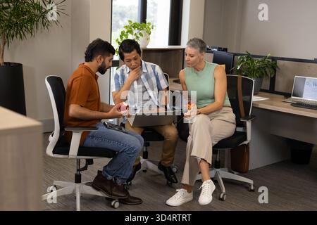 Discutere di diversi colleghi seduti intorno a un notebook in un moderno ufficio open space, con mele rosse Foto Stock