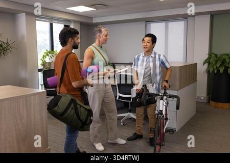 Parla di diversi colleghi in piedi alla reception dell'ufficio, con bicicletta, tappetino per lo yoga e tazza di caffè Foto Stock