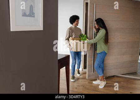 Diverse amiche che si scambiano scatole di cartone nell'ingresso di casa moderna, con piante in vaso Foto Stock