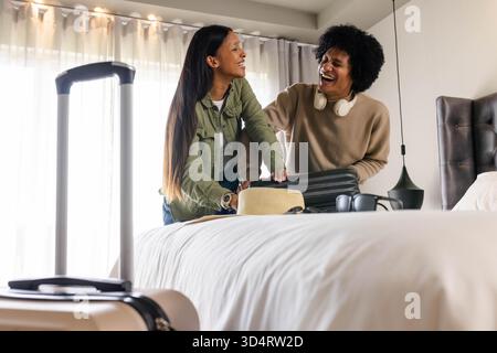 Una coppia che si diverte a ridere e che prepara una valigia nera arrotolata sul letto in camera d'albergo, con un cappello di paglia Foto Stock