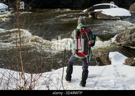 Giovane uomo adulto caucasico in piedi sulla riva innevata del fiume tenendo in mano pali da trekking e indossando un grande zaino da trekking, di fronte al fiume che scorre con rocce innevate sullo sfondo Foto Stock